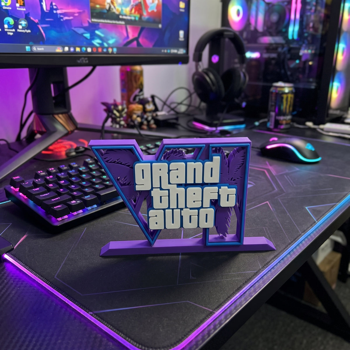 A 3D-printed Grand Theft Auto V desk stand featuring the game's logo and silhouettes of Liberty City, displayed on a gaming desk with a keyboard, mouse, and monitor in a room setup.