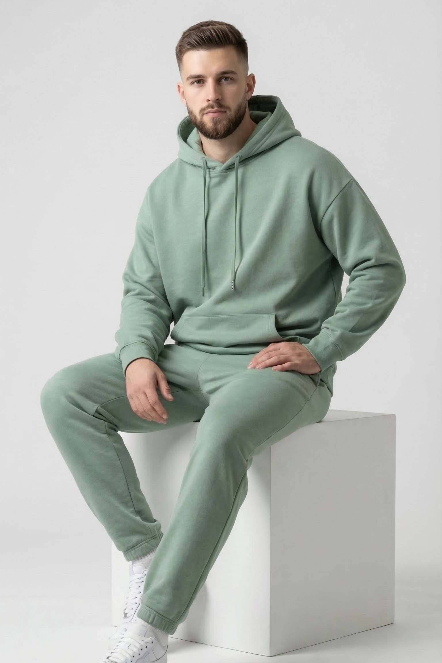 Man wearing a light green hoodie and pants sitting on a white cube against a light gray background
