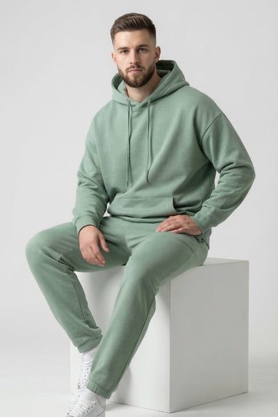 Man wearing a light green hoodie and pants sitting on a white cube against a light gray background