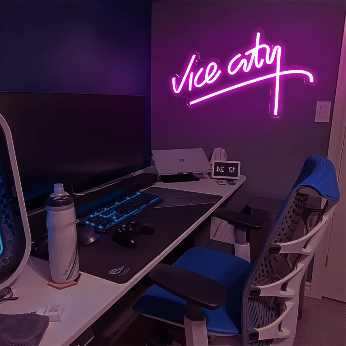 Neon 'Vice City' sign above a desk with computer equipment in a dark room.