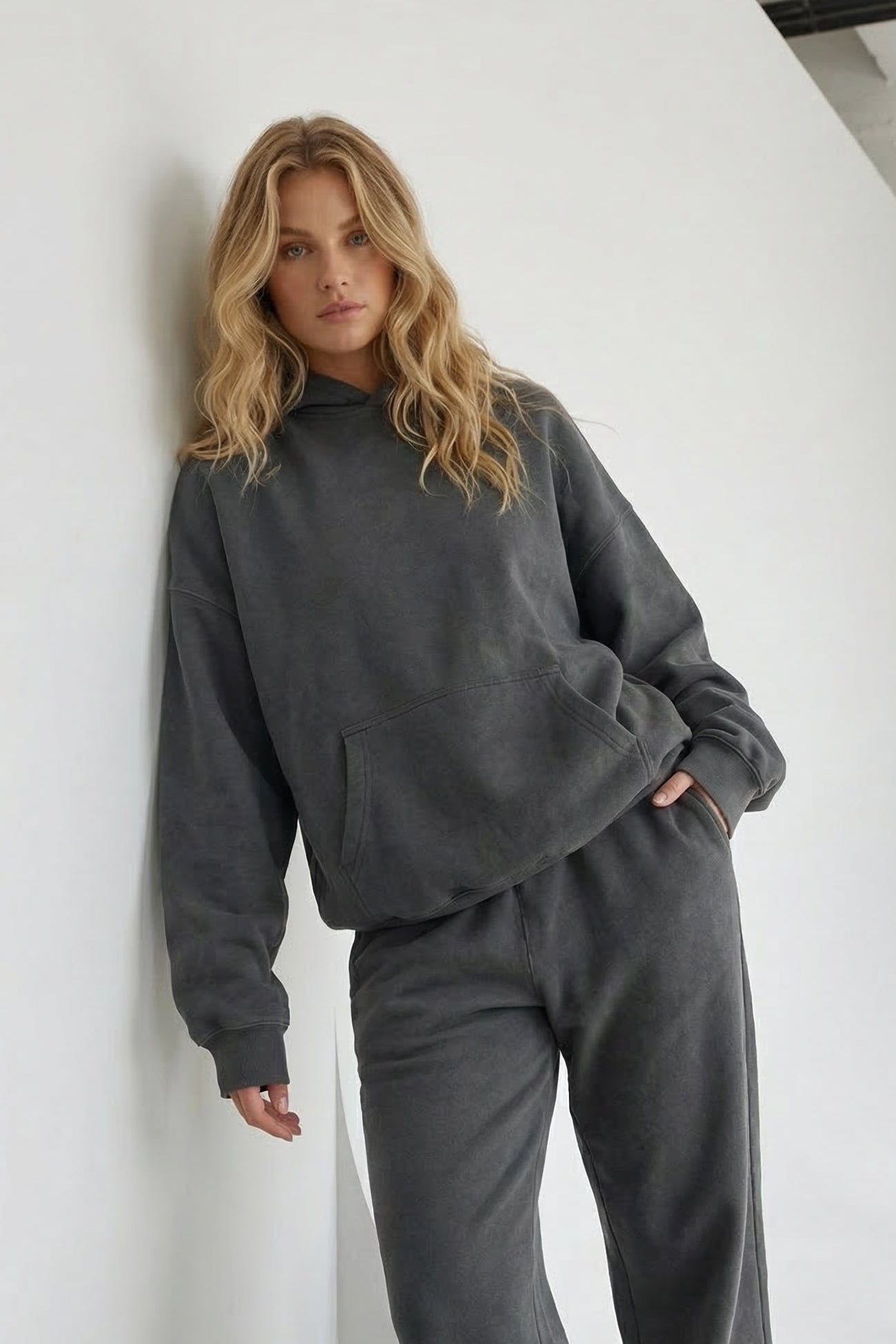 Woman wearing a gray tracksuit leaning against a white wall.