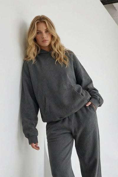 Woman wearing a gray tracksuit leaning against a white wall.