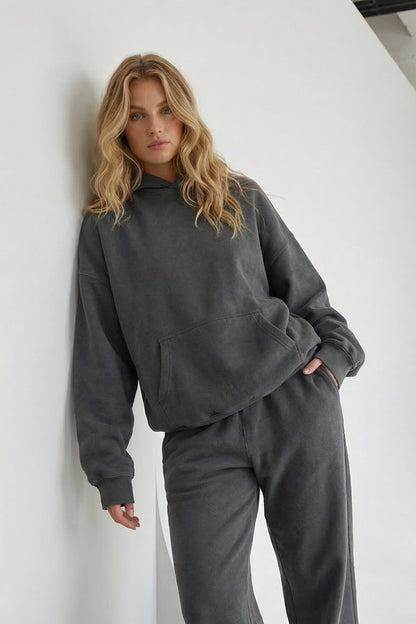 Woman wearing a gray tracksuit leaning against a white wall.