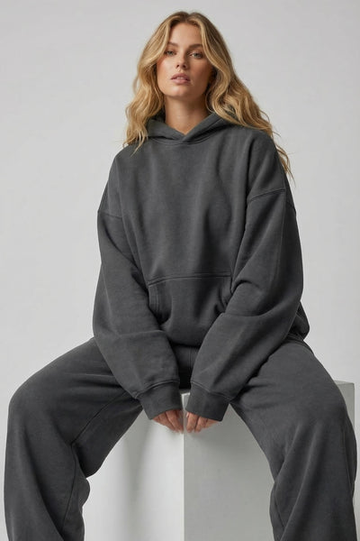 Person wearing a dark gray hoodie and pants sitting on a white stool against a light gray background