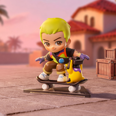 A stylized collectible figure of a character with bright yellow hair riding a skateboard in a desert town setting. The figure wears a multicolor outfit with orange, yellow, black, and purple details, set against a softly blurred background of buildings and sand.