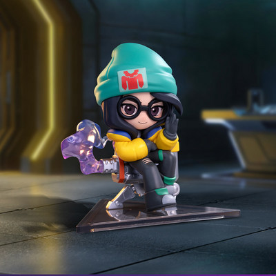 Killjoy A stylized chibi figurine of a female character in a teal beanie with a red logo, black glasses, a yellow jacket, and gray pants, posed on a clear display base with a translucent purple weapon accessory in a dark futuristic interior.