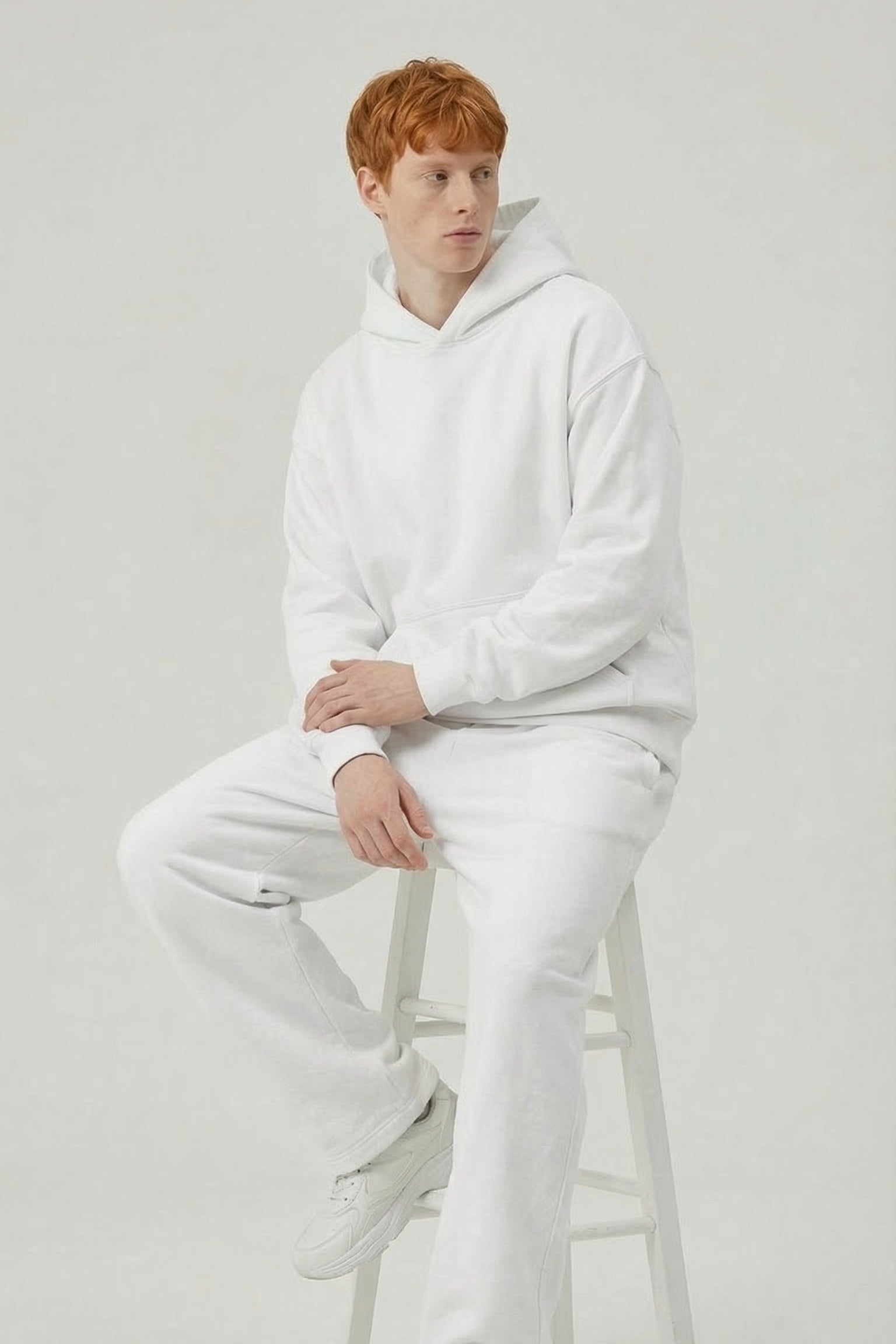 Person wearing a white hoodie and pants sitting on a stool against a plain background