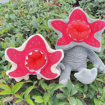 Gray plush character doll with red star hood sitting among green leaves